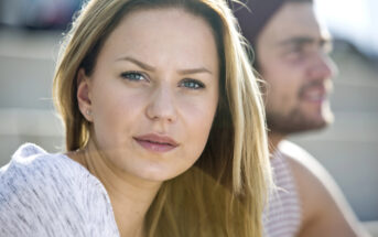 A woman with long blonde hair and blue eyes looks directly at the camera with a neutral expression. She is wearing a light-colored top. In the background, there is a man with a beard and a hat, slightly out of focus, gazing into the distance.