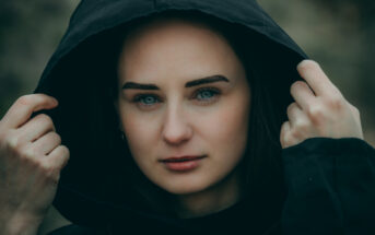 A person with light skin and dark hair is looking directly at the camera while holding the hood of a black hoodie with both hands. The background is blurred, drawing attention to the person's face.