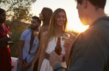 A group of young adults is enjoying an outdoor gathering at sunset, holding drinks and smiling. The warm light of the setting sun illuminates their faces as they converse. Trees and a scenic landscape are visible in the background.