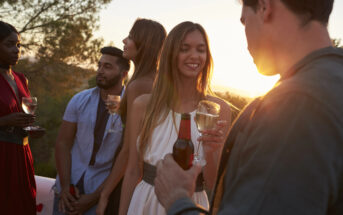 A group of young adults is enjoying an outdoor gathering at sunset, holding drinks and smiling. The warm light of the setting sun illuminates their faces as they converse. Trees and a scenic landscape are visible in the background.