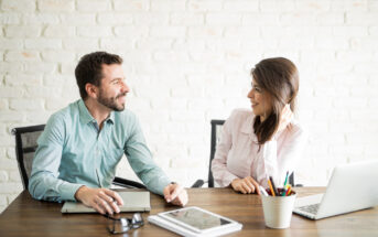 A man and a woman are sitting at a wooden table in a modern office setting. They are smiling and engaged in a conversation. On the table are a laptop, a tablet, a notebook, glasses, and a cup holding various pens and pencils.