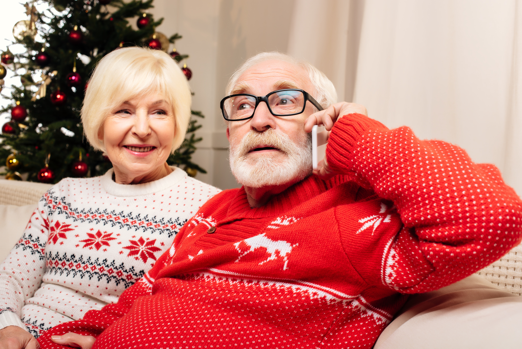 An elderly couple in festive sweaters sits on a couch in front of a decorated Christmas tree. The man, wearing glasses and a red sweater with reindeer, is talking on the phone. The woman, in a white sweater with red snowflakes, is smiling at him.