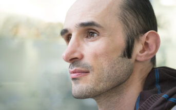 A man with a shaved head on top and short dark hair on the sides is looking to the left. He has a trimmed beard and is wearing a brown jacket with a blue stripe on the shoulder. The background is blurred and light-colored.