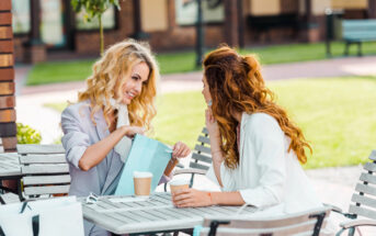 Two women with curly hair wearing light-colored outfits sit at an outdoor café table with several shopping bags. One woman excitedly shows the other a blue bag, while both hold paper coffee cups. A green lawn and buildings are visible in the background.