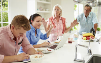 A family of four gathers in a bright kitchen. A young man and woman on the left are focused on a tablet and laptop respectively. An older woman in the center appears to be talking passionately, while an older man on the right listens intently. The counter has breakfast items.