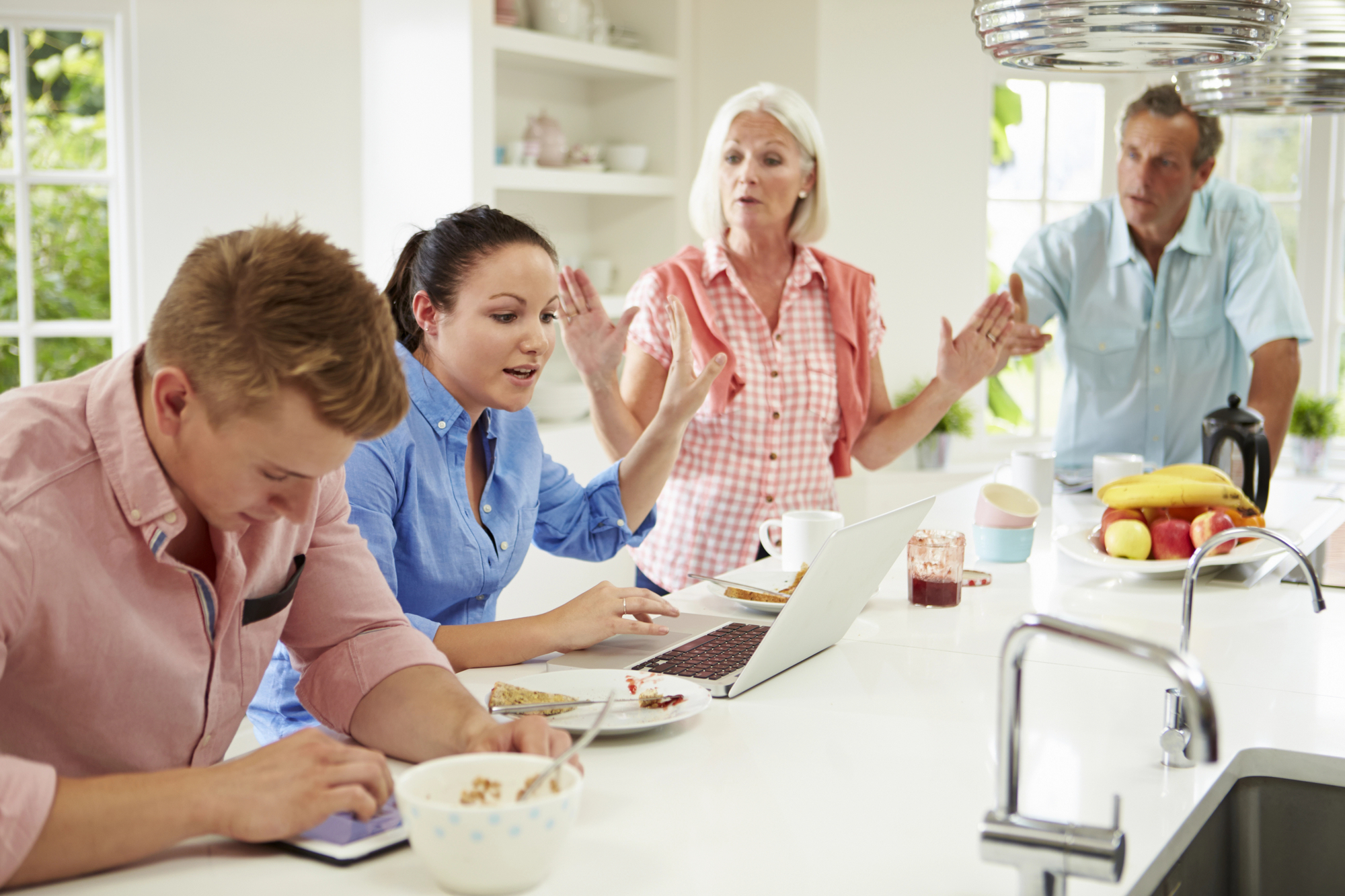 A family of four gathers in a bright kitchen. A young man and woman on the left are focused on a tablet and laptop respectively. An older woman in the center appears to be talking passionately, while an older man on the right listens intently. The counter has breakfast items.