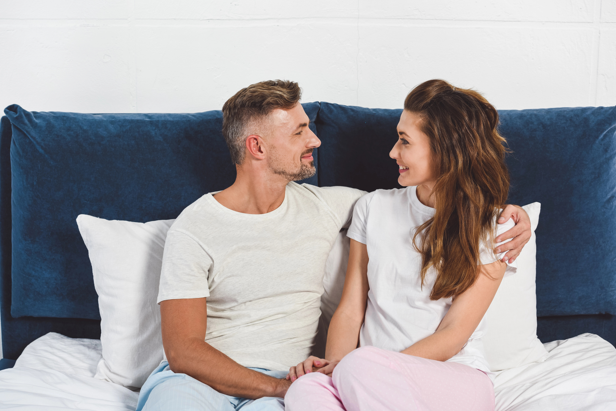 A man and a woman sit on a bed with a dark blue headboard, facing and smiling at each other. Both are wearing casual clothes; the man is in a white t-shirt, and the woman is in a white top. The bedding is white and they appear relaxed and happy.