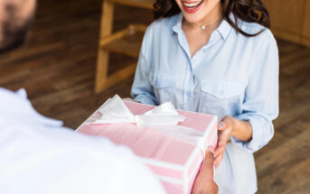 A person with long dark hair and a light blue shirt smiles while receiving a wrapped pink gift box with a white ribbon from another person. The image captures a moment of joy and exchange of a present indoors.