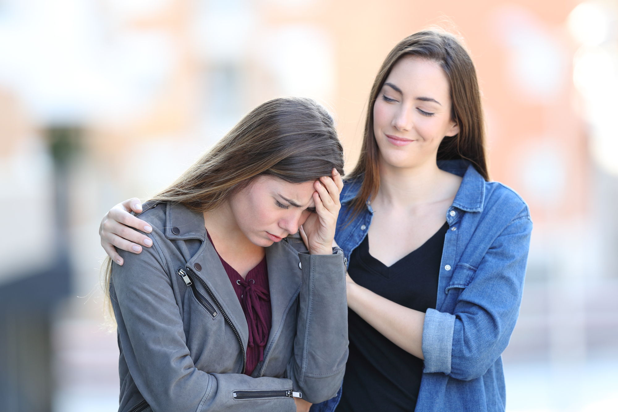 Two women are outdoors. One woman looks distressed, holding her head with one hand and her other arm crossed. The other woman, wearing a denim shirt, stands beside her with one hand on her shoulder and a reassuring smile, offering comfort.