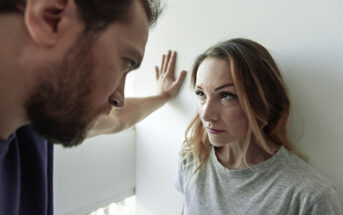 A man stands close to a woman, with his arm raised and hand against the wall, looking down at her. The woman looks up at him with a serious expression. The scene suggests a tense or confrontational moment.