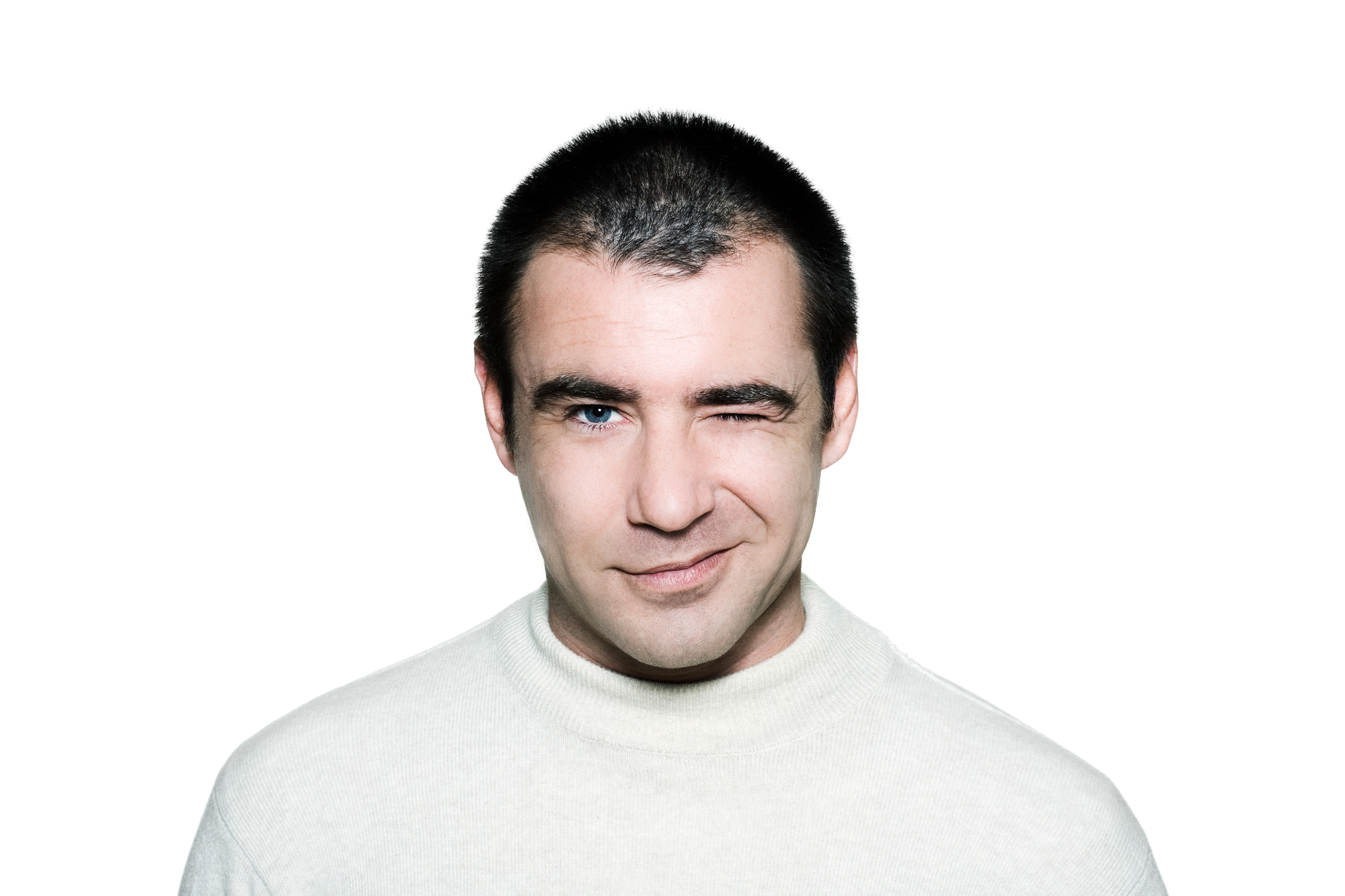 A man with short dark hair wearing a white turtleneck sweater is standing against a plain white background. He is smiling slightly and has one eye closed, as if winking.