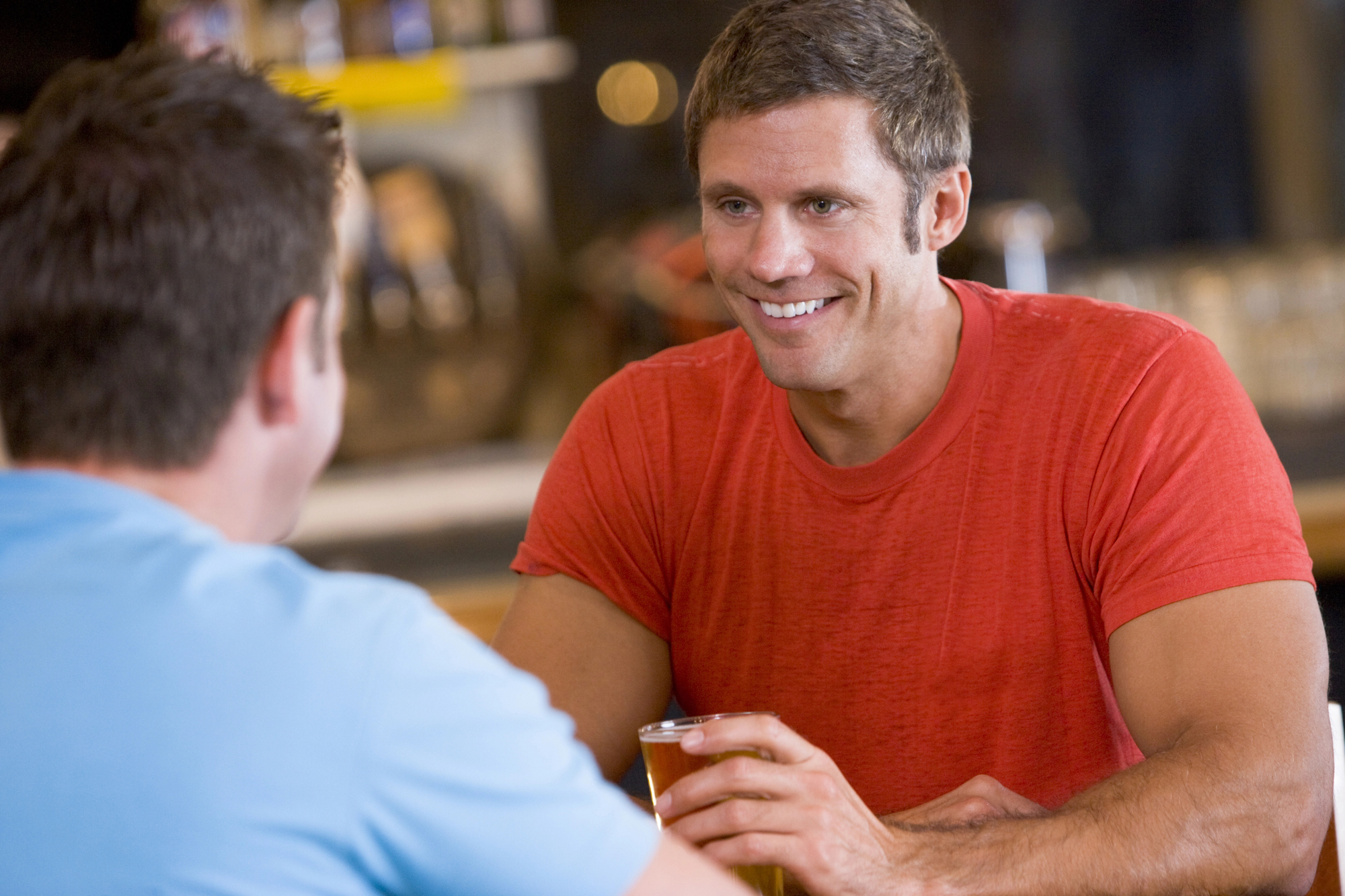 Two men having a conversation in a casual indoor setting. One man, in a blue shirt, faces away from the camera, while the man in a red shirt smiles and holds a drink, looking at the other with an engaging expression. The background is slightly blurred.