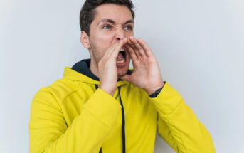 A man wearing a bright yellow hoodie with a black hood is standing against a plain white background. He is shouting with his hands cupped around his mouth. His expression is intense, and he appears to be calling out or yelling with emotion.