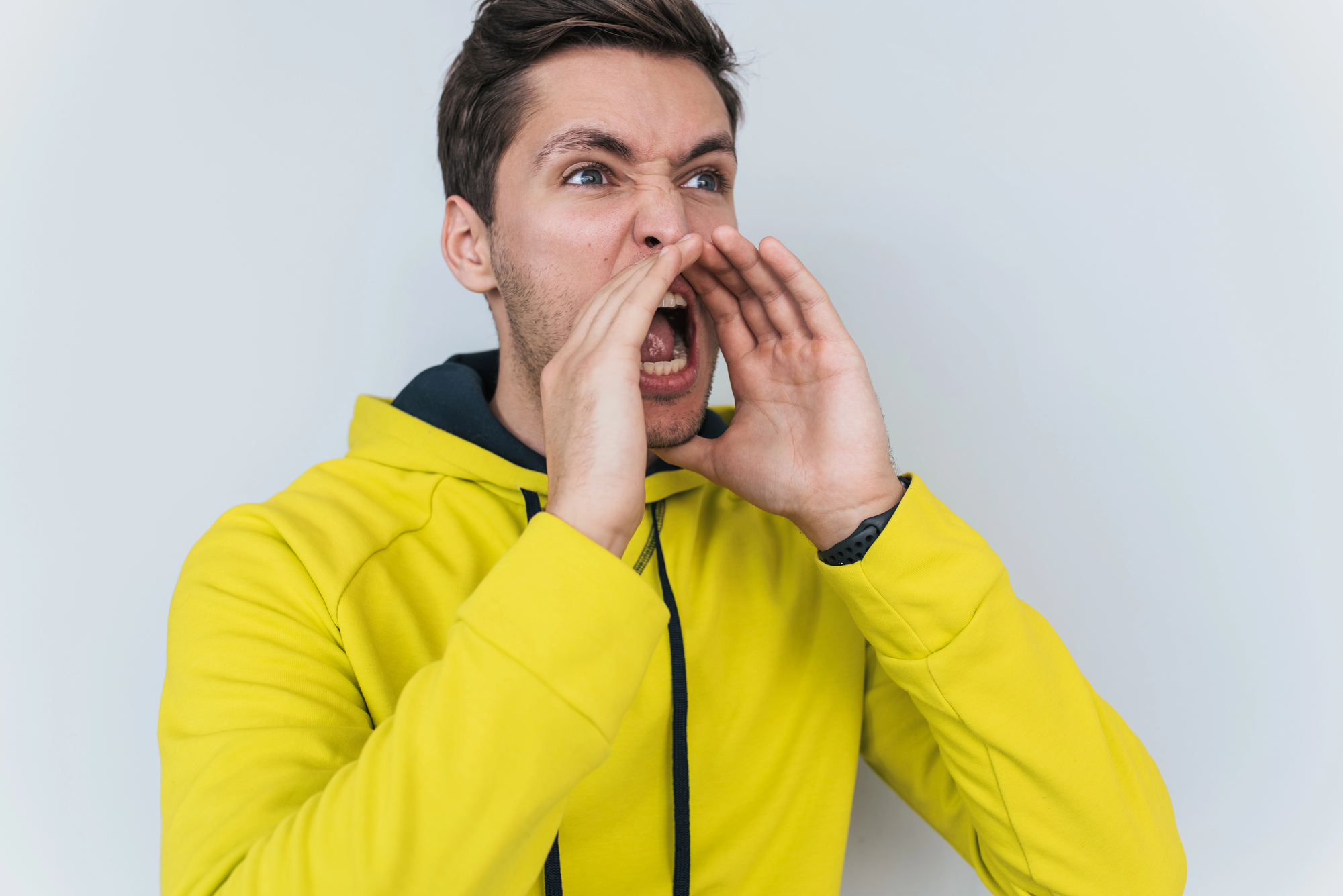 A man wearing a bright yellow hoodie with a black hood is standing against a plain white background. He is shouting with his hands cupped around his mouth. His expression is intense, and he appears to be calling out or yelling with emotion.