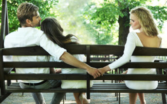 A man, seated on a park bench, embraces a woman with long brown hair resting on his shoulder while holding hands behind her back with a blonde woman in a white dress, seated to his other side. Sunlight filters through green trees in the background.