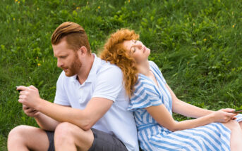 A man and a woman are sitting back-to-back on a grassy field. The man, with a serious expression, is focused on his smartphone, while the woman, wearing a striped dress, leans her head back with her eyes closed, looking relaxed.