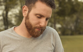 A man with a beard, short hair, and ear gauges is looking down. He is wearing a light gray T-shirt and standing outdoors on a green grassy area, with trees visible in the blurred background. The lighting suggests it's a calm, possibly cloudy day.