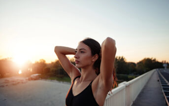 A woman with long hair stands on a bridge at sunset, with her arms raised and hands resting on the back of her head. She is dressed in a black sleeveless top and gazes thoughtfully into the distance. The sky is clear, and the sun is low on the horizon.