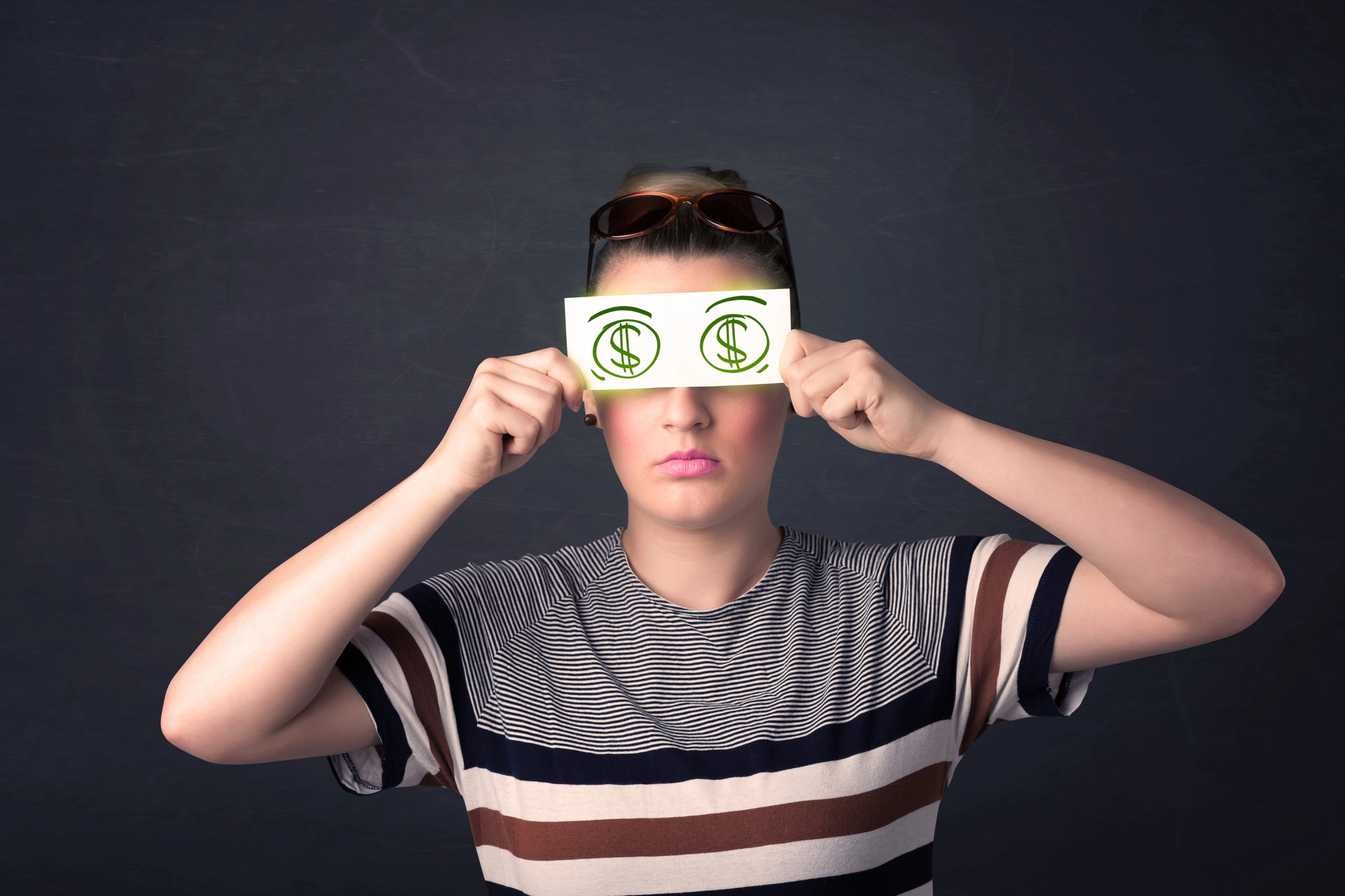 A person wearing a striped shirt and sunglasses on their head holds up a piece of paper in front of their eyes. The paper has green dollar signs drawn on it, making it appear as though they have dollar sign eyes. The background is dark and plain.