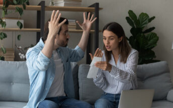 A man and woman are sitting on a couch in a living room, engaged in a heated discussion. The man has his hands raised in frustration, while the woman is holding a piece of paper and gesturing as she talks. Shelves with books and plants are in the background.