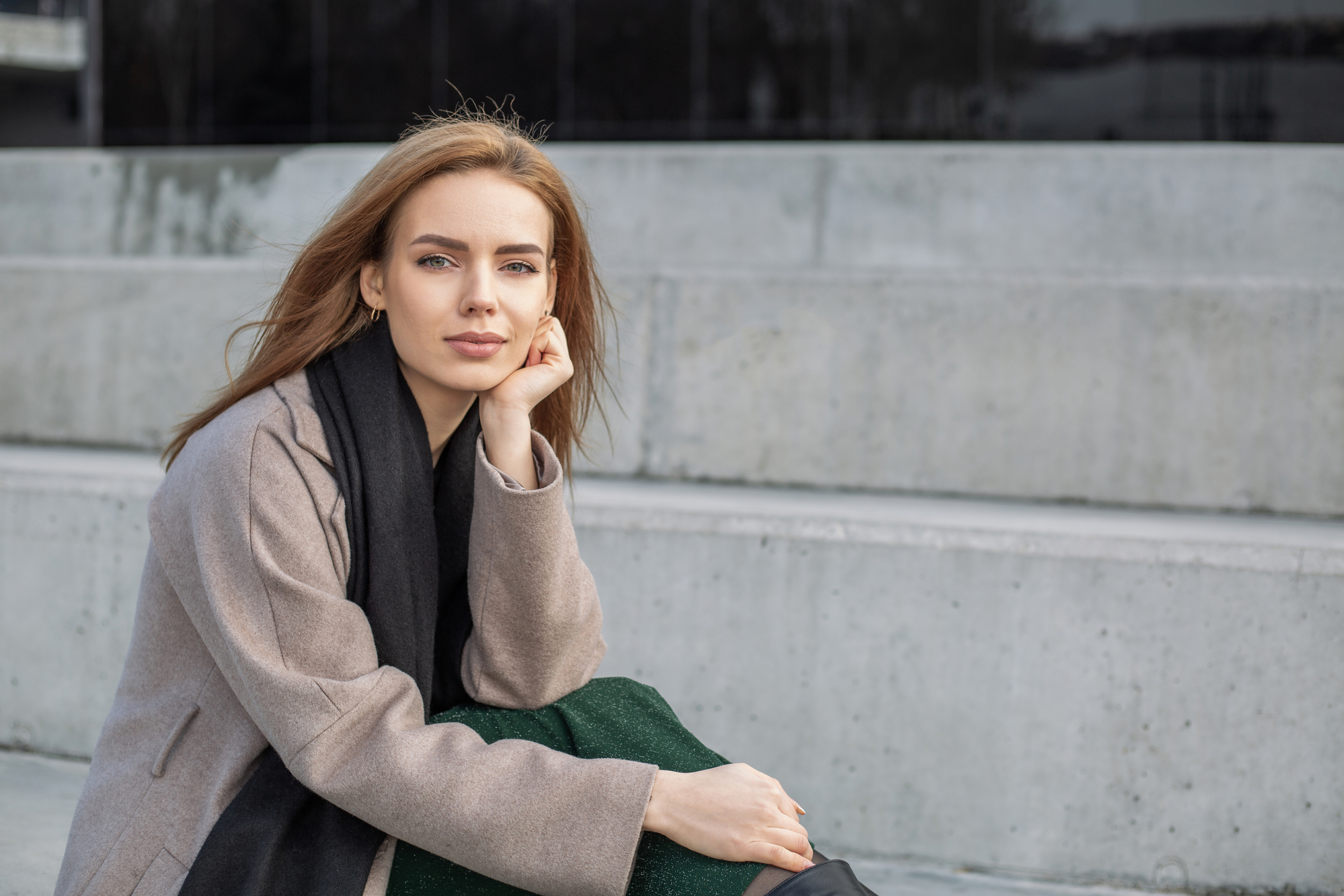 A woman with light brown hair wearing a beige coat and black scarf sits on concrete steps, resting her face on her hand and looking towards the camera with a neutral expression.