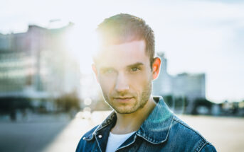 A man with short hair stands outside in a city setting, wearing a denim jacket. The sun is shining brightly behind him, creating a lens flare effect. The background includes blurred buildings and a partly cloudy sky.