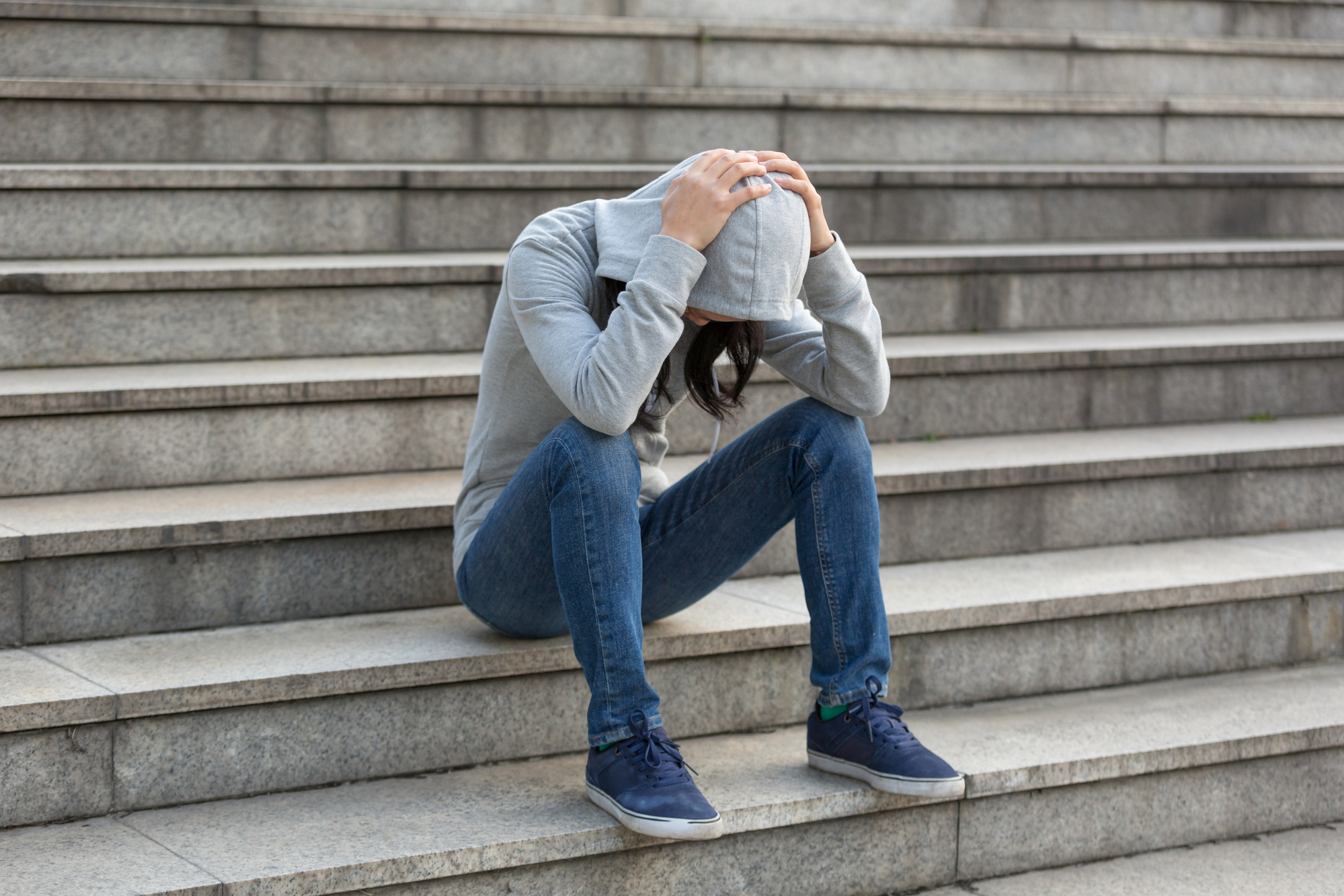 A person wearing a gray hoodie and blue jeans sits on stone steps with their head down, hands clasping the top of their head. The person appears to be in distress or deep thought.