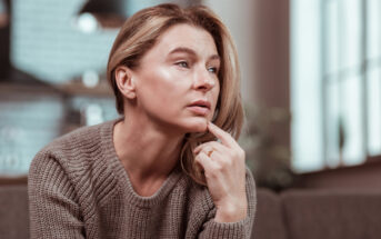 A woman with light hair sits pensively on a couch, resting her chin on her hand. She is wearing a light brown sweater and looking slightly to the side. The background is softly blurred, showing a cozy indoor setting with natural light coming through the windows.