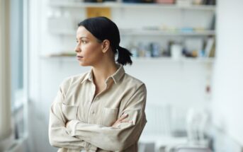 A woman with her hair tied back is standing indoors with her arms crossed, looking thoughtfully out of the window. She is wearing a beige button-up shirt. There are shelves with various items and books blurred in the background.