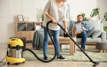 A woman in a casual outfit is vacuuming the floor with a yellow and silver vacuum cleaner in a living room. In the background, a man in a denim jacket and jeans is sitting on a light blue sofa, eating snacks and watching her. The room is modern and bright.