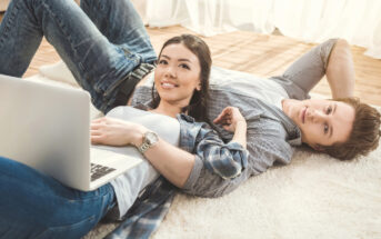 A young woman and man are laying on a carpeted floor with a laptop in front of them. The woman is smiling and looking up, while the man lies beside her with one arm behind his head, gazing ahead. Both are casually dressed in jeans and shirts.