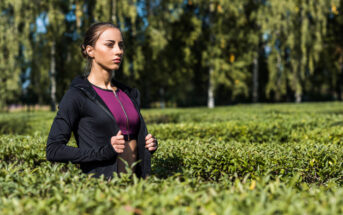 A woman stands in a field of shrubs, zipping up her black hoodie over a purple top. She looks into the distance with a serious expression. The background features green trees with bright sunlight filtering through.