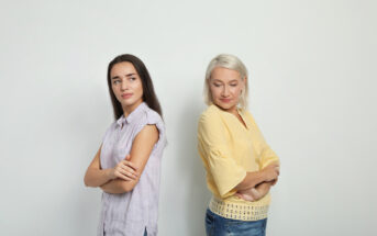 Two women, one with long dark hair and the other with short white hair, stand back-to-back with their arms crossed, both looking slightly annoyed or displeased. They are dressed casually in light-colored tops and blue jeans, standing against a plain white background.