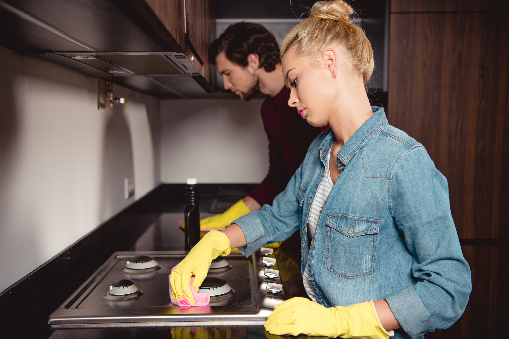 A woman and a man, both wearing yellow rubber gloves, are cleaning the kitchen. The woman in focus is scrubbing the stove with a pink cloth while the man in the background is wiping a surface with a yellow cloth. They are working in a modern kitchen with wooden cabinets.