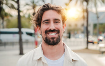 A man with medium-length hair and a beard is smiling outdoors. He is wearing a light-colored jacket and a white shirt. The background features palm trees and the sun setting, creating a warm, pleasant atmosphere.