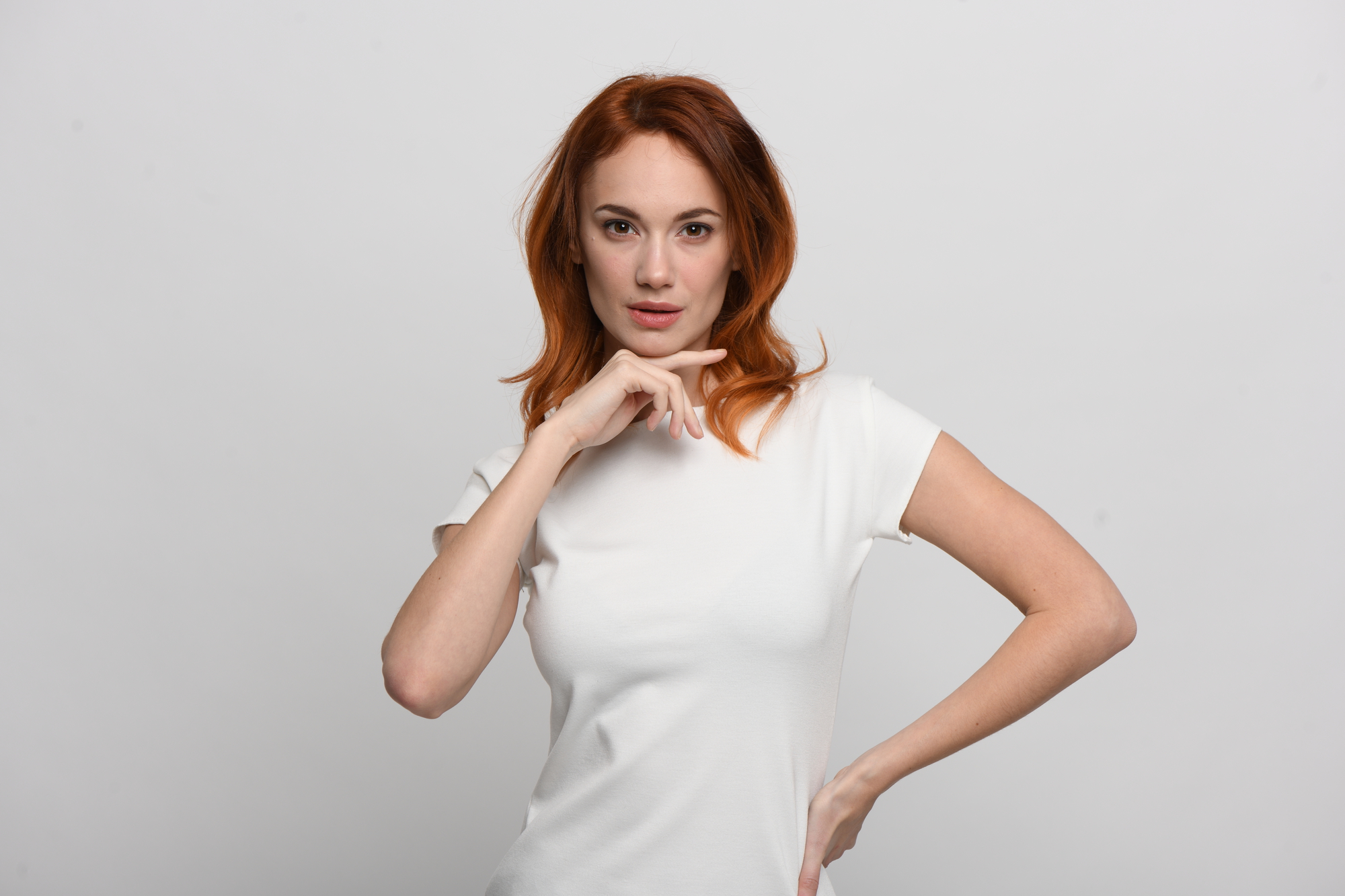 A woman with red hair stands against a plain gray background. She is wearing a white short-sleeved shirt and has a calm expression. One hand is placed on her hip, and the other hand is raised with her fingers touching her chin.