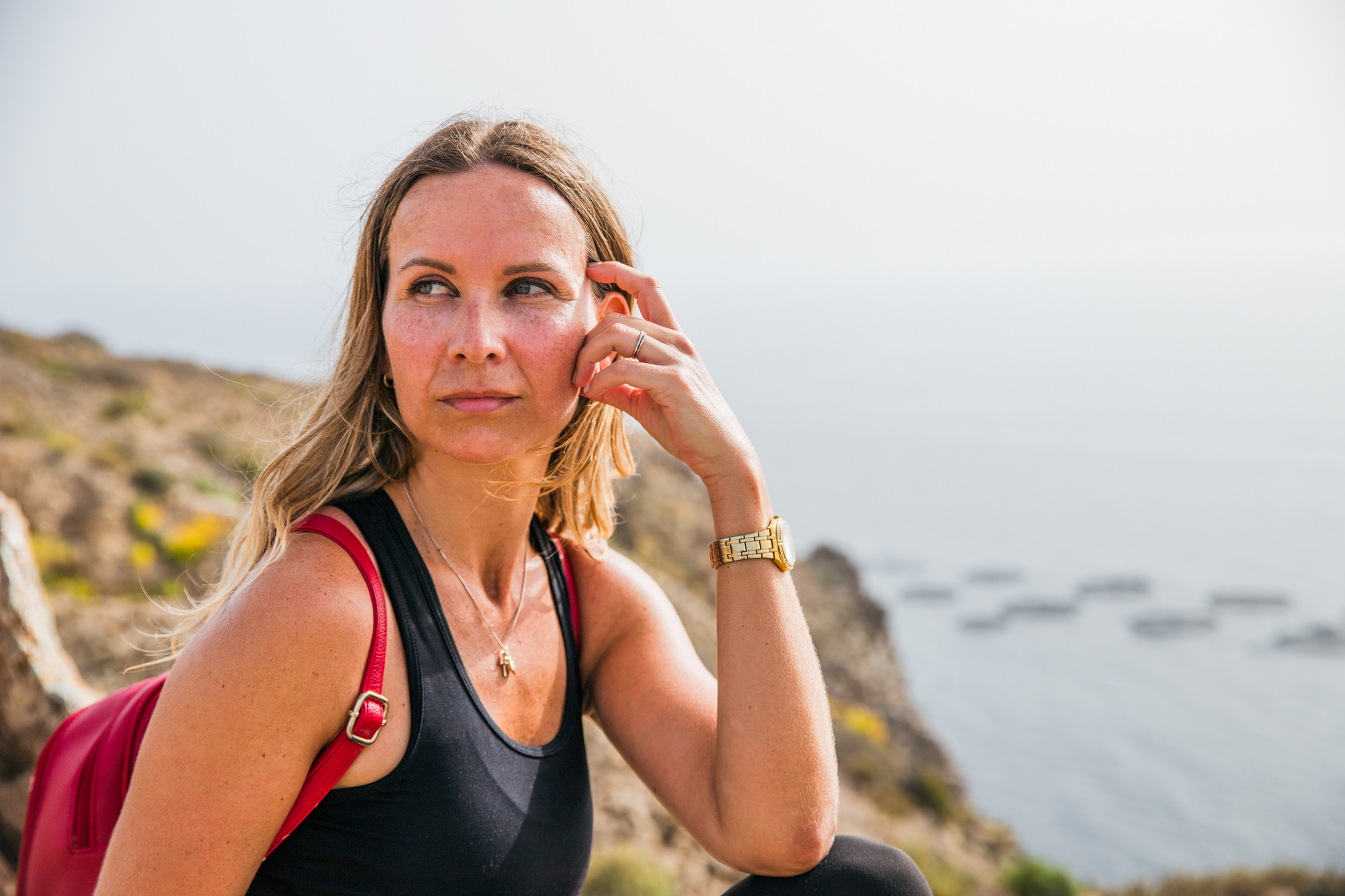 A woman with blonde hair wearing a black tank top and red backpack sits thoughtfully on a rocky cliff overlooking the ocean. She rests her chin on her hand, gazing into the distance. The ocean and a hazy sky are visible in the background.