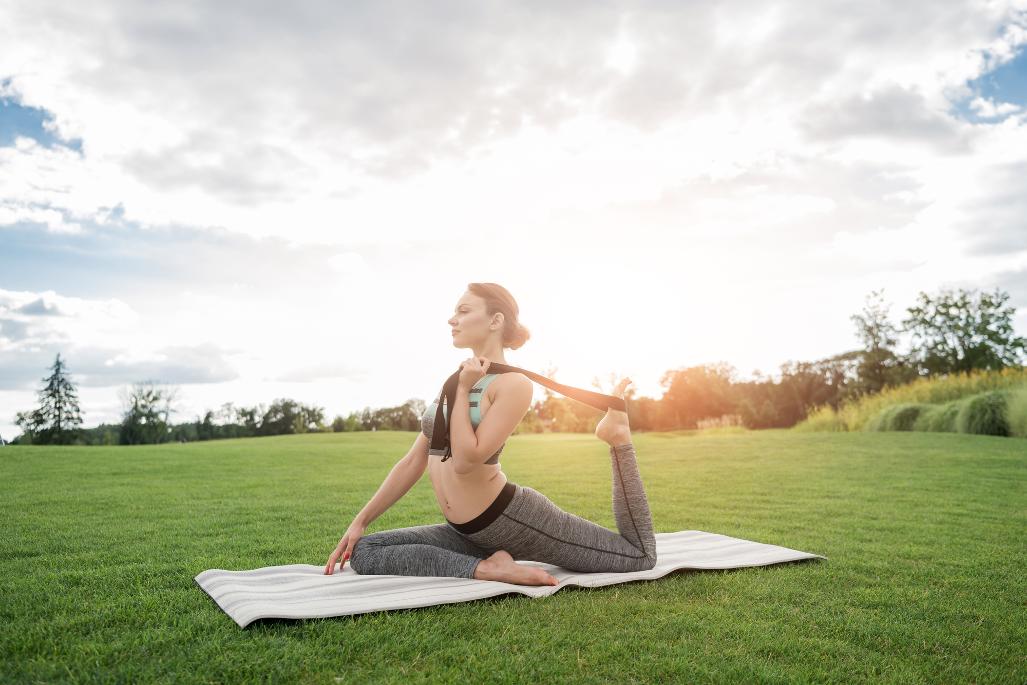 A person practices yoga on a mat in a grassy field under a partly cloudy sky. They are in a pose with one knee bent forward and the other leg extended backward, holding the foot with a strap over their shoulder, facing towards the sun which is setting or rising in the background.