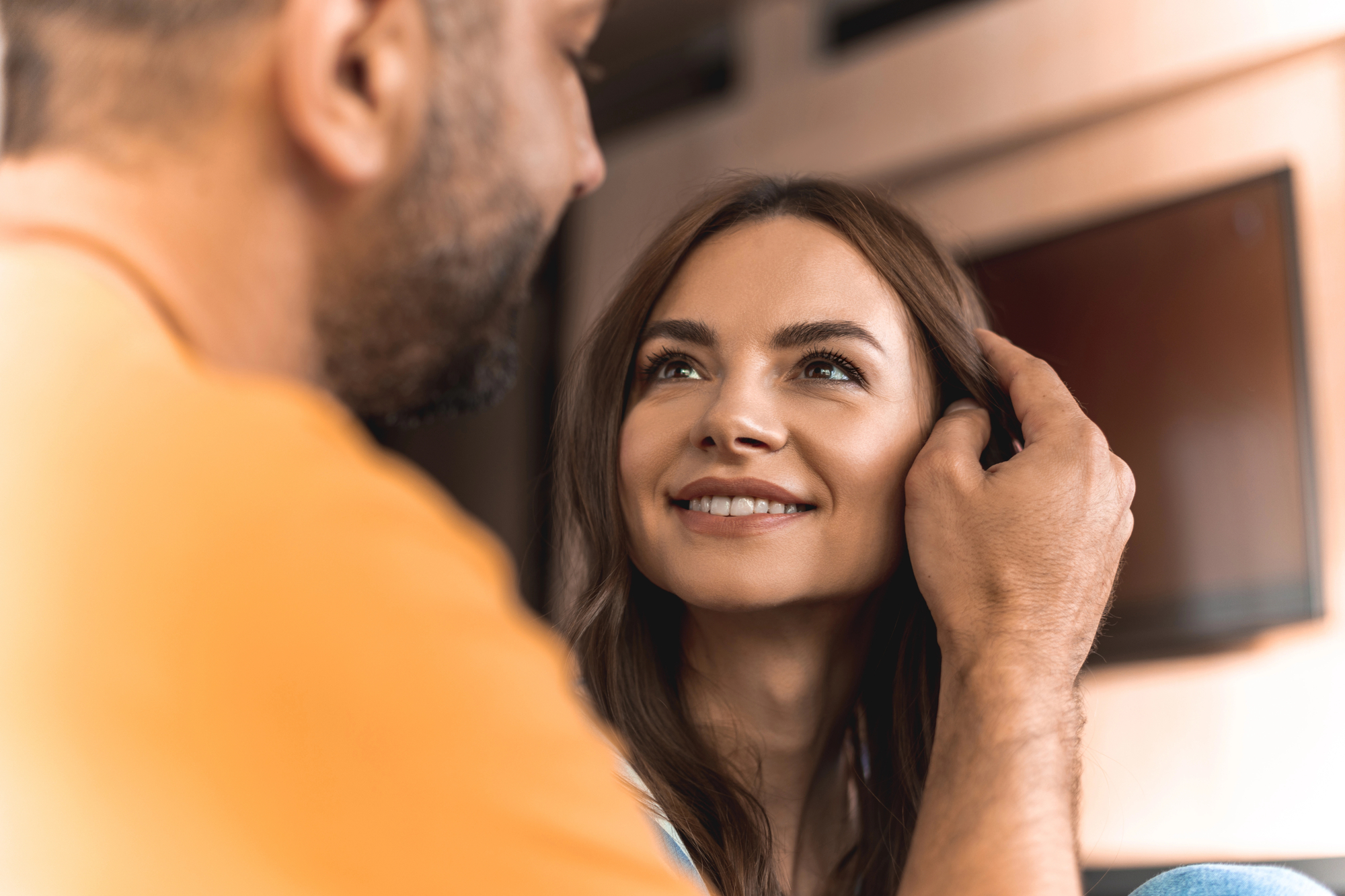 A woman with long brown hair smiles warmly, looking up at a man who is gently touching her hair. The scene appears to be indoors, with a blurred background including a wall-mounted television. The lighting is soft and warm.