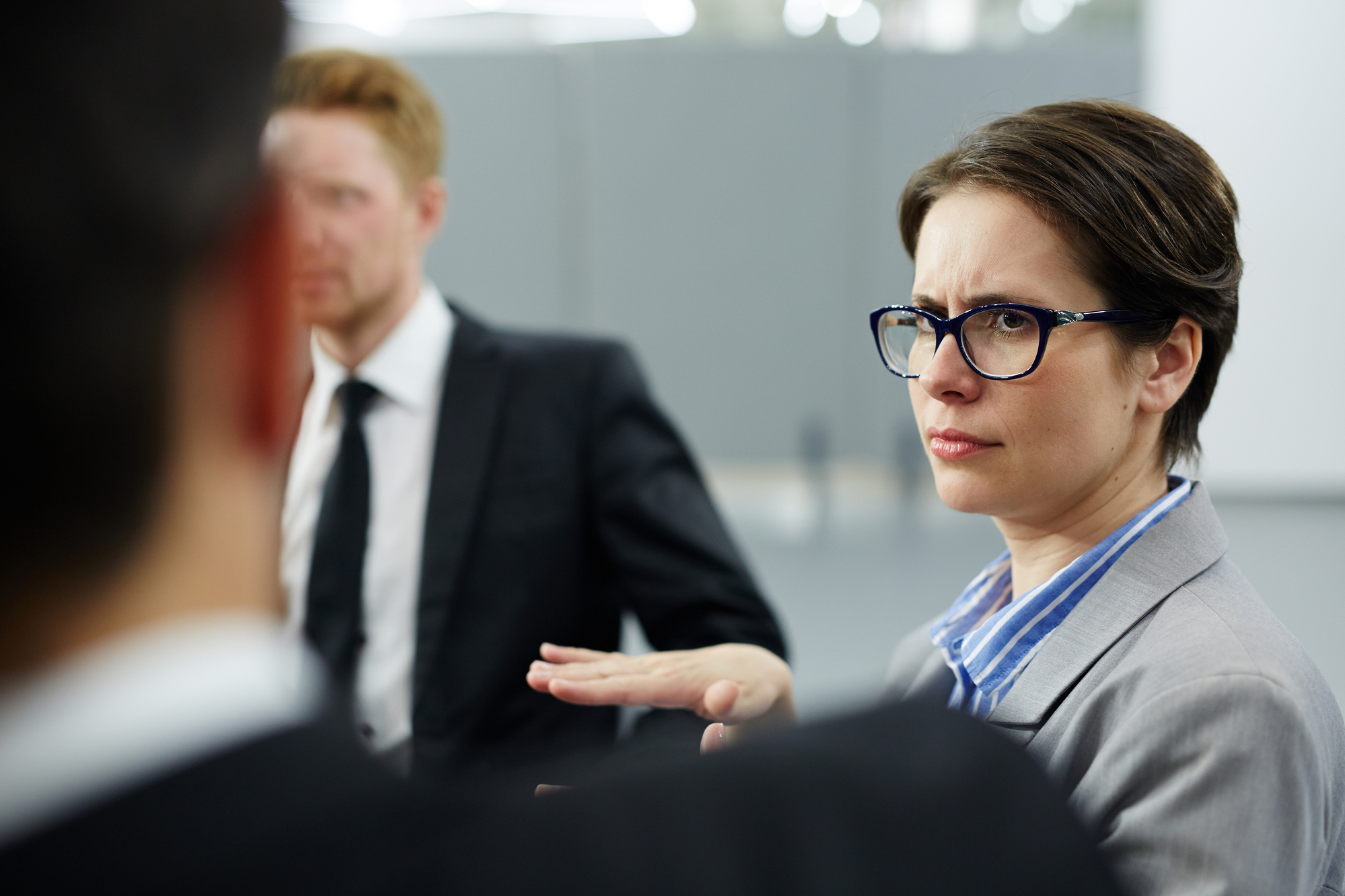 A woman wearing glasses and a gray suit is engaged in a serious conversation with two men, both dressed in business attire. Her hand is slightly raised as she listens intently. They are in an office setting with gray partitions in the background.