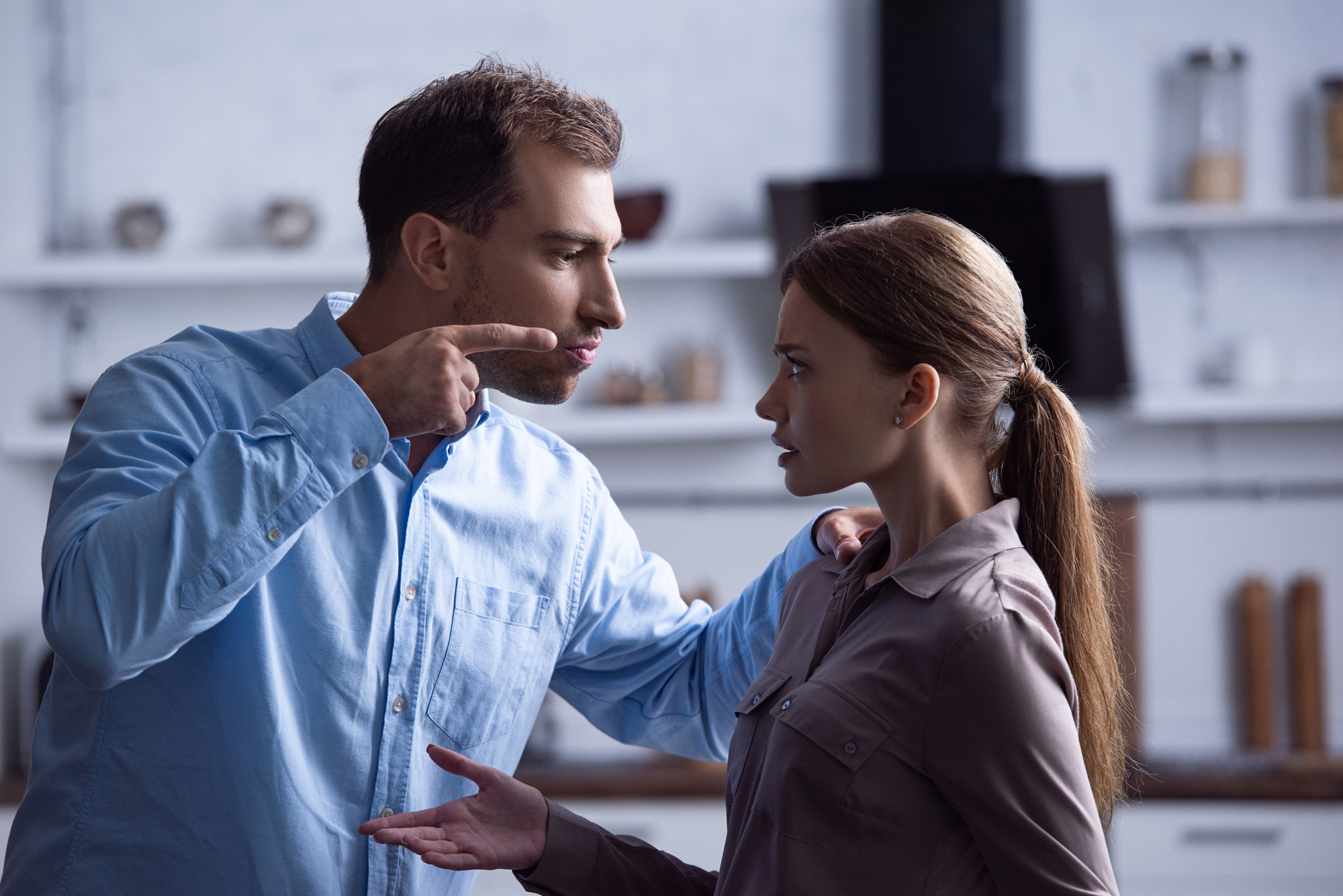 A man and a woman are having an intense argument in a kitchen. The man, wearing a light blue shirt, is pointing his finger at the woman, who is wearing a gray blouse. Both appear upset, with the woman gesturing with her hands. Kitchen items are blurred in the background.