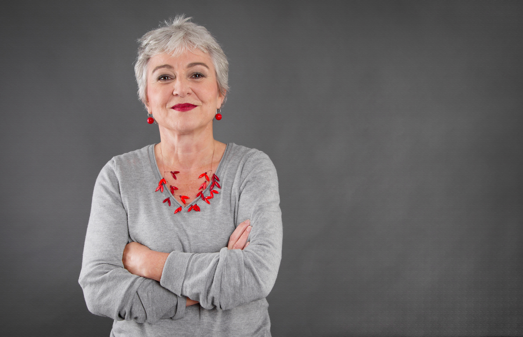 An older woman with short gray hair stands against a gray background, smiling confidently with her arms crossed. She is wearing a gray sweater, red earrings, and a matching red necklace.