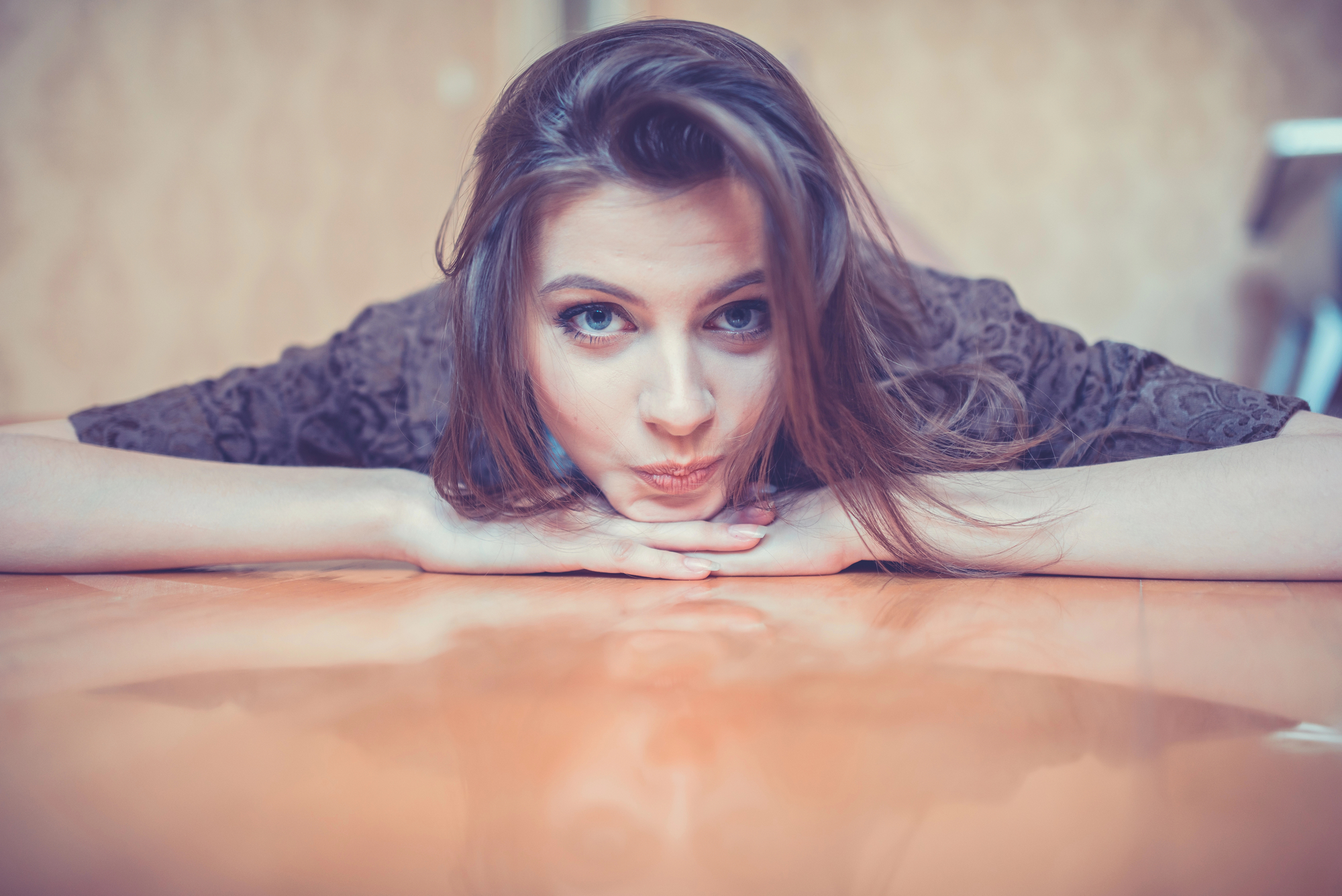 A young person with long brown hair lies face down on a shiny wooden floor, resting their chin on their hands. They have a playful expression with their eyes looking directly at the camera. The background is softly blurred.