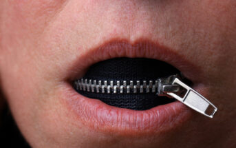 Close-up of a person's lips partially unzipped with a zipper running horizontally across their mouth, revealing black fabric behind the zipper. The image suggests the concept of silence or keeping quiet.