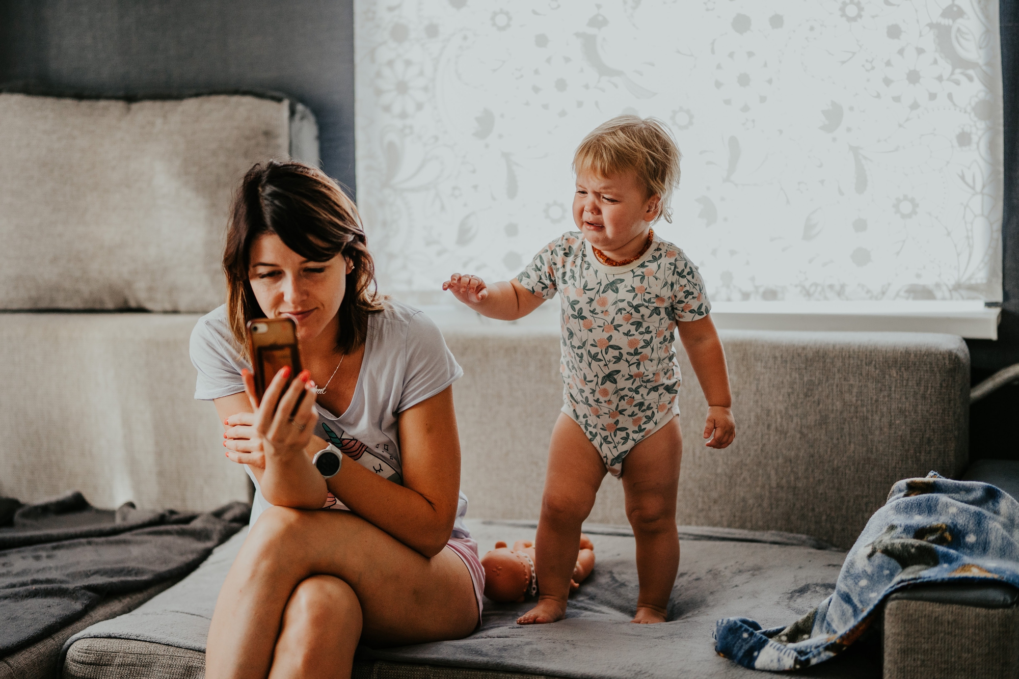 A woman sitting on a couch looks at her phone while a toddler standing next to her cries and reaches out. The toddler is wearing a patterned onesie. There are toys and a blanket on the couch, and a bright window with a floral-patterned curtain in the background.