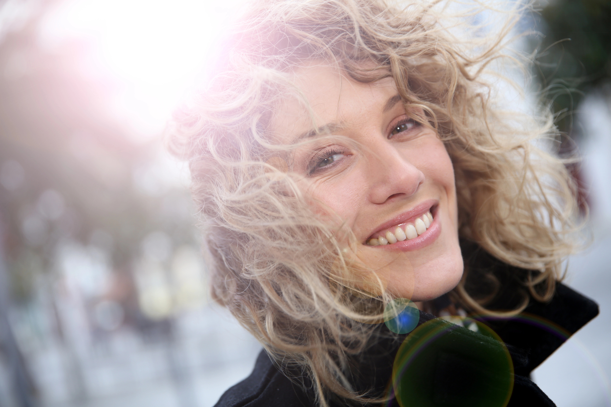 A person with curly blond hair smiles brightly while looking at the camera. The background is softly blurred, and sunlight creates a halo effect around their hair, contributing to the cheerful and warm atmosphere of the photo.