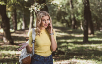 A woman with blonde hair, wearing a yellow shirt and carrying a backpack, stands in a forested area talking on her phone. She looks confused or frustrated, with one hand raised in an expressive gesture. The lush green background suggests it's a sunny day.