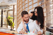 In a contemporary office setting, a man in a white shirt and blue tie sits at a desk looking at papers. A woman with long dark hair and glasses stands behind him, leaning over his shoulder, looking at the papers too. The office has a modern, airy feel with large windows.