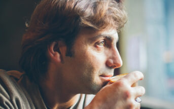 A thoughtful person with wavy, light brown hair gently holds a cup close to their mouth while looking out into the distance. They are wearing a casual shirt, and soft daylight illuminates their face, highlighting a serene and contemplative expression.