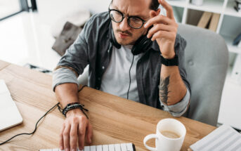 A man wearing glasses and headphones sits at a desk with a MIDI keyboard, working intently. His left hand is on the keyboard while his right hand is raised, appearing thoughtful. On the desk are a laptop, a cup of coffee, and a notepad.